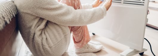 Using heater at home in winter. Woman warming her hands sitting by device and wearing warm clothes. Heating season.